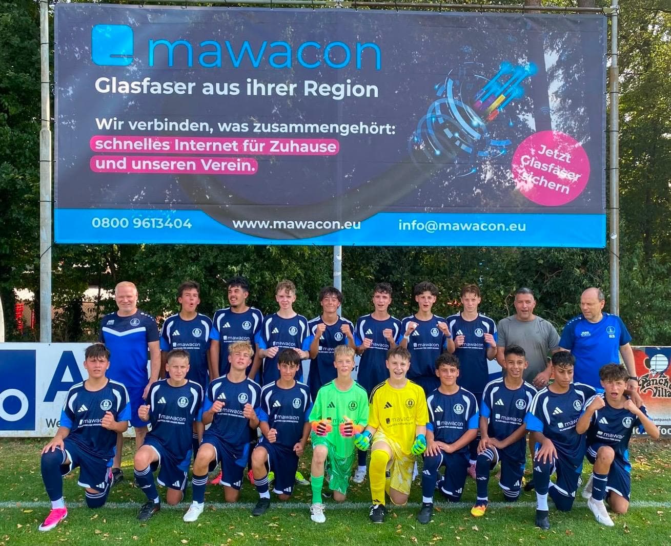 Jugendmannschaft in dunkelblauen Trikots mit Mawacon-Aufdruck vor einer Bandenwerbung mit der Aufschrift „Glasfaser aus ihrer Region“ auf dem Sportplatz in Rodenbach